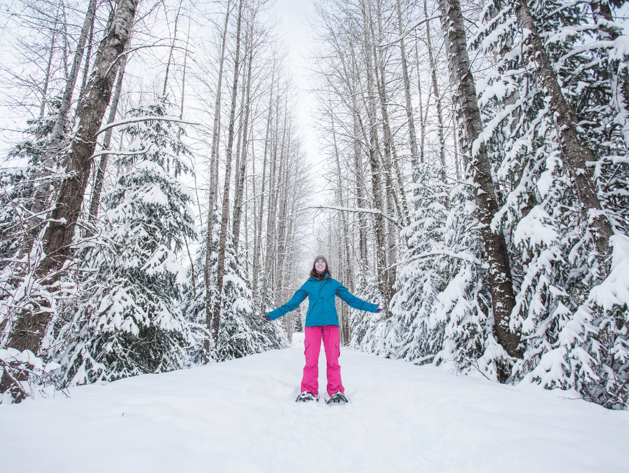 A woman in pink snow pants stands with open arms amidst snowy trees, conveying the joy of winter adventures and scoring affordable gear.