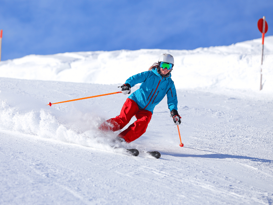 A skier in red pants and a blue jacket carves through fresh powder on a sunny slope.