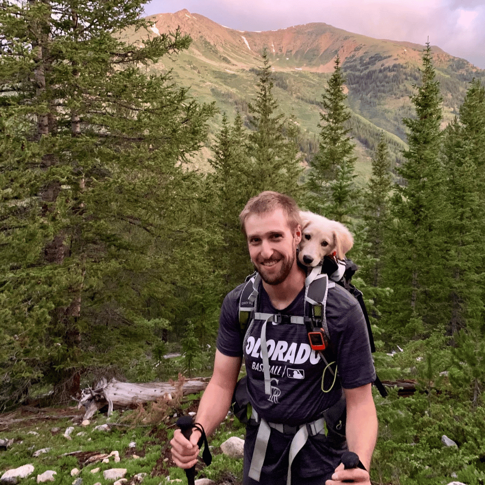Brian smiles, carrying a cute puppy in a backpack while hiking amidst tall Colorado pines and mountains.