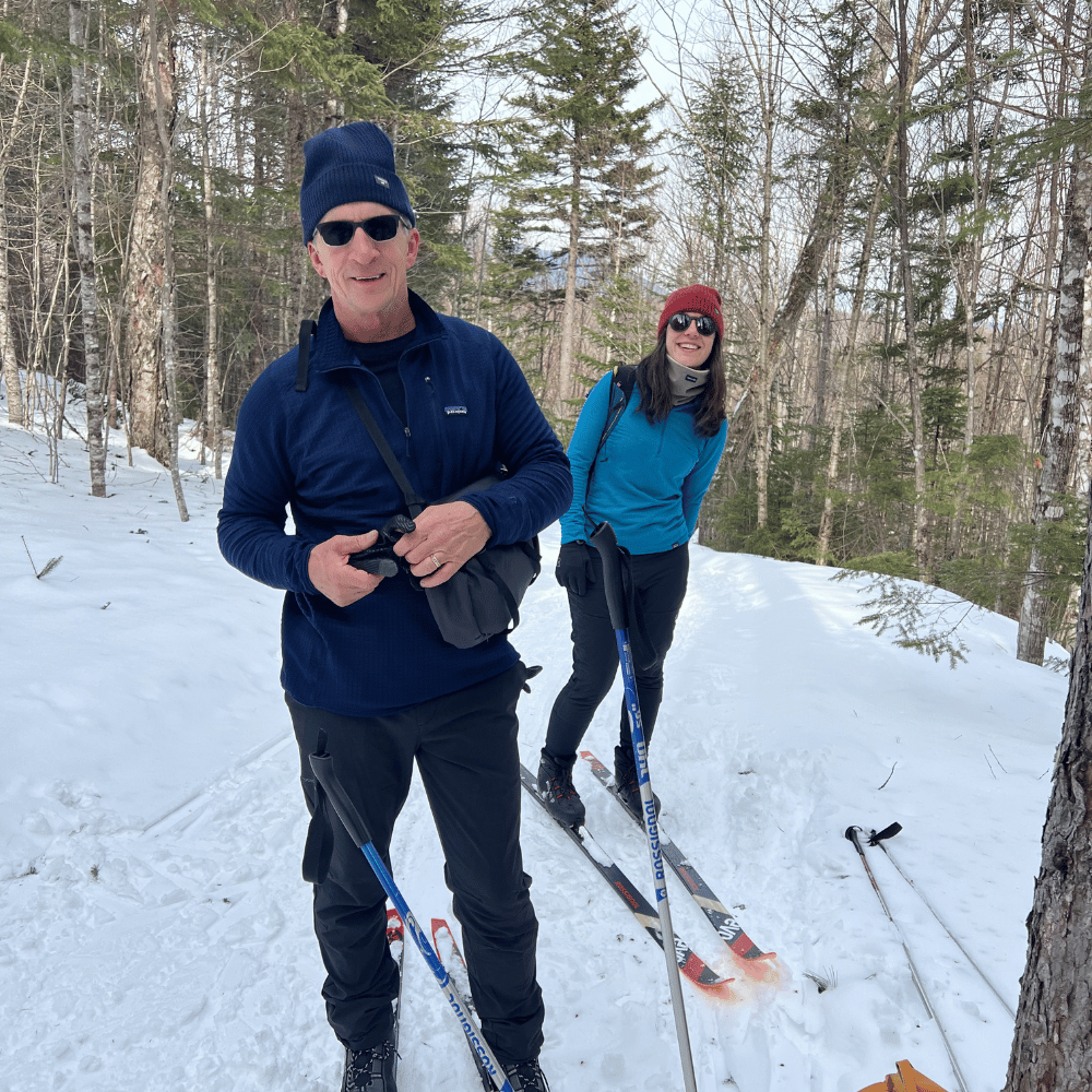 David Cuccaro and a woman smiling while cross-country skiing on a snowy trail through a forest.