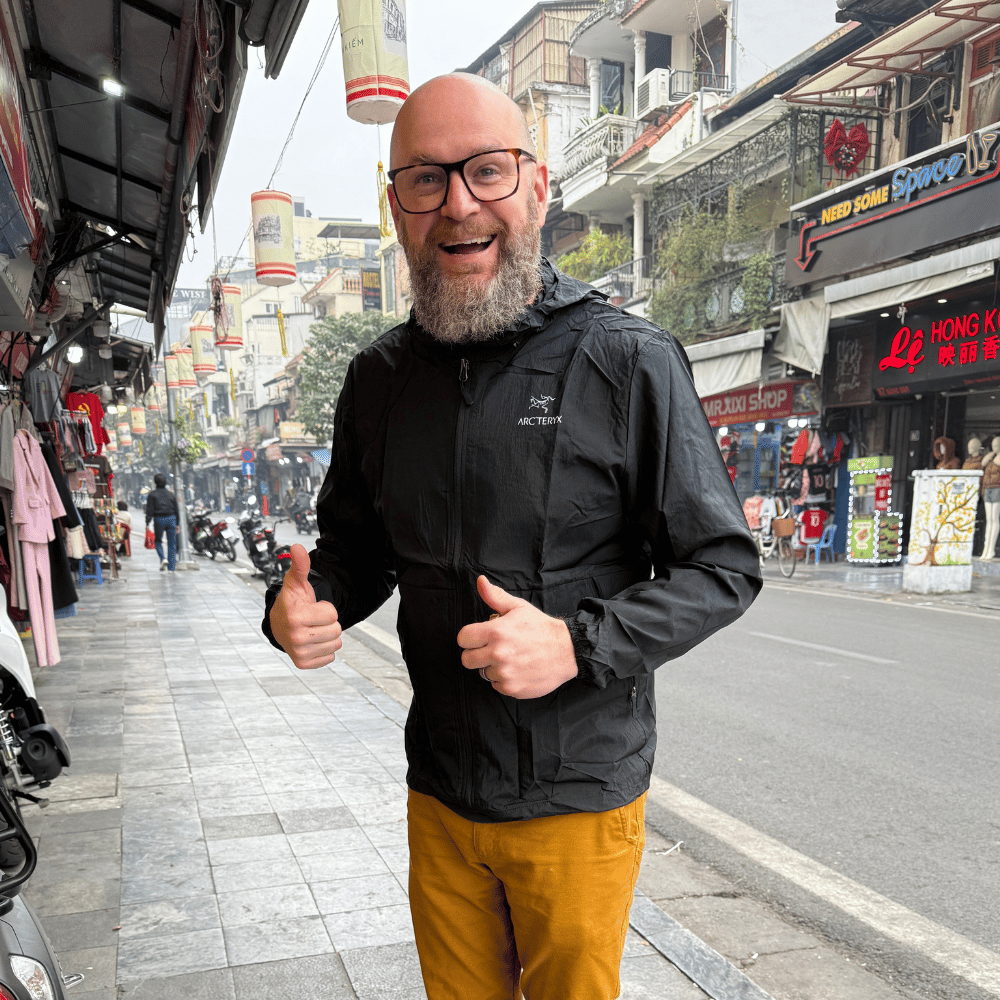Jimmy Funkhouser, thrilled in Hanoi, gives two thumbs up. His black jacket contrasts with mustard pants against a busy street backdrop.