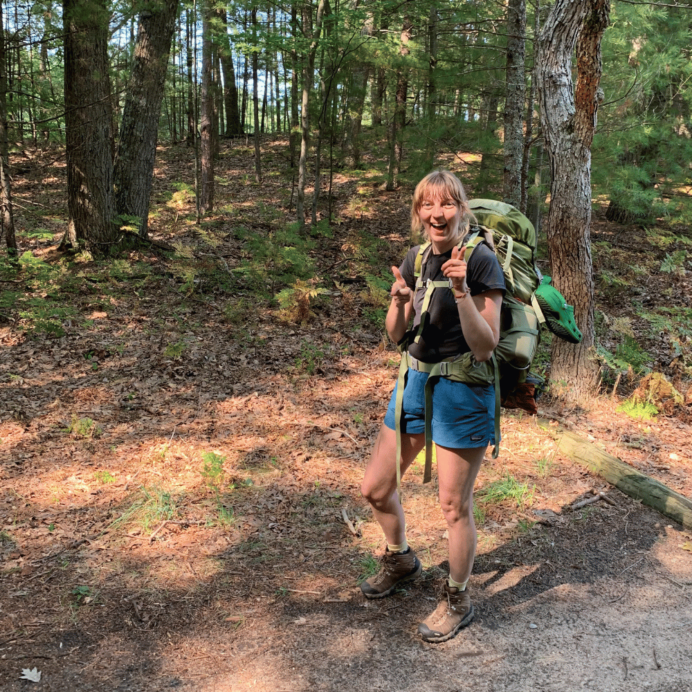 Enthusiastic Liz Lane gives two thumbs up on a sun-dappled trail, her backpacking pack promising adventure.