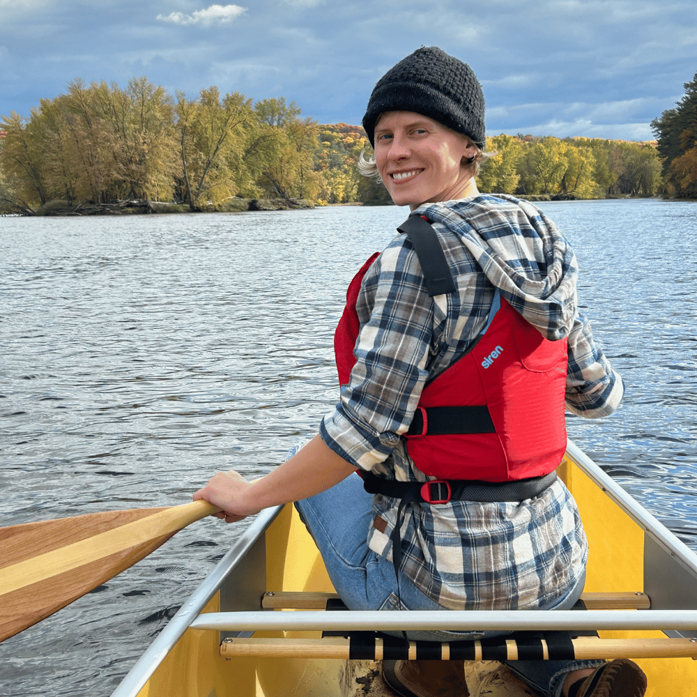 A smiling Whitney paddles a yellow canoe on calm water, surrounded by autumn foliage, exuding peaceful adventure.