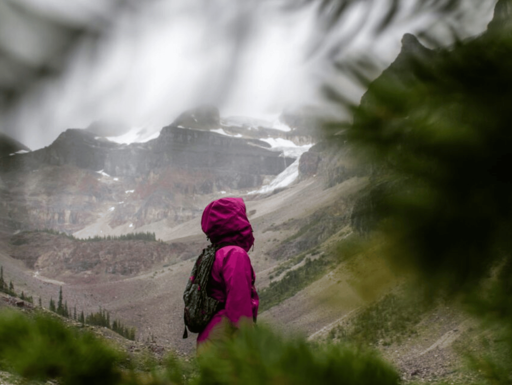 A lone hiker in a magenta jacket gazes at a majestic, misty mountain range, evoking a sense of adventure and awe.