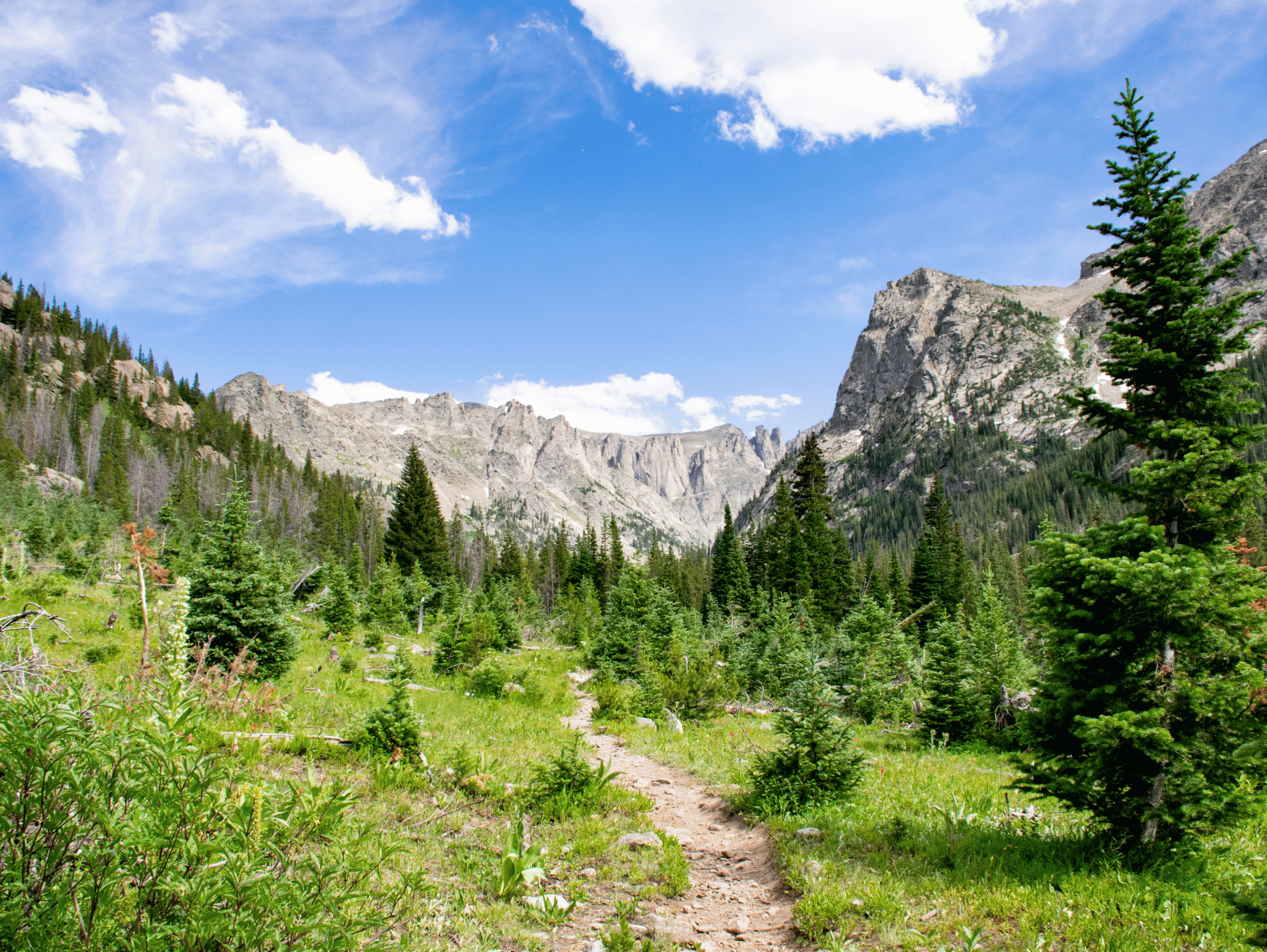 A dirt path winds through a vibrant green meadow towards jagged mountain peaks, inviting adventure and exploration.