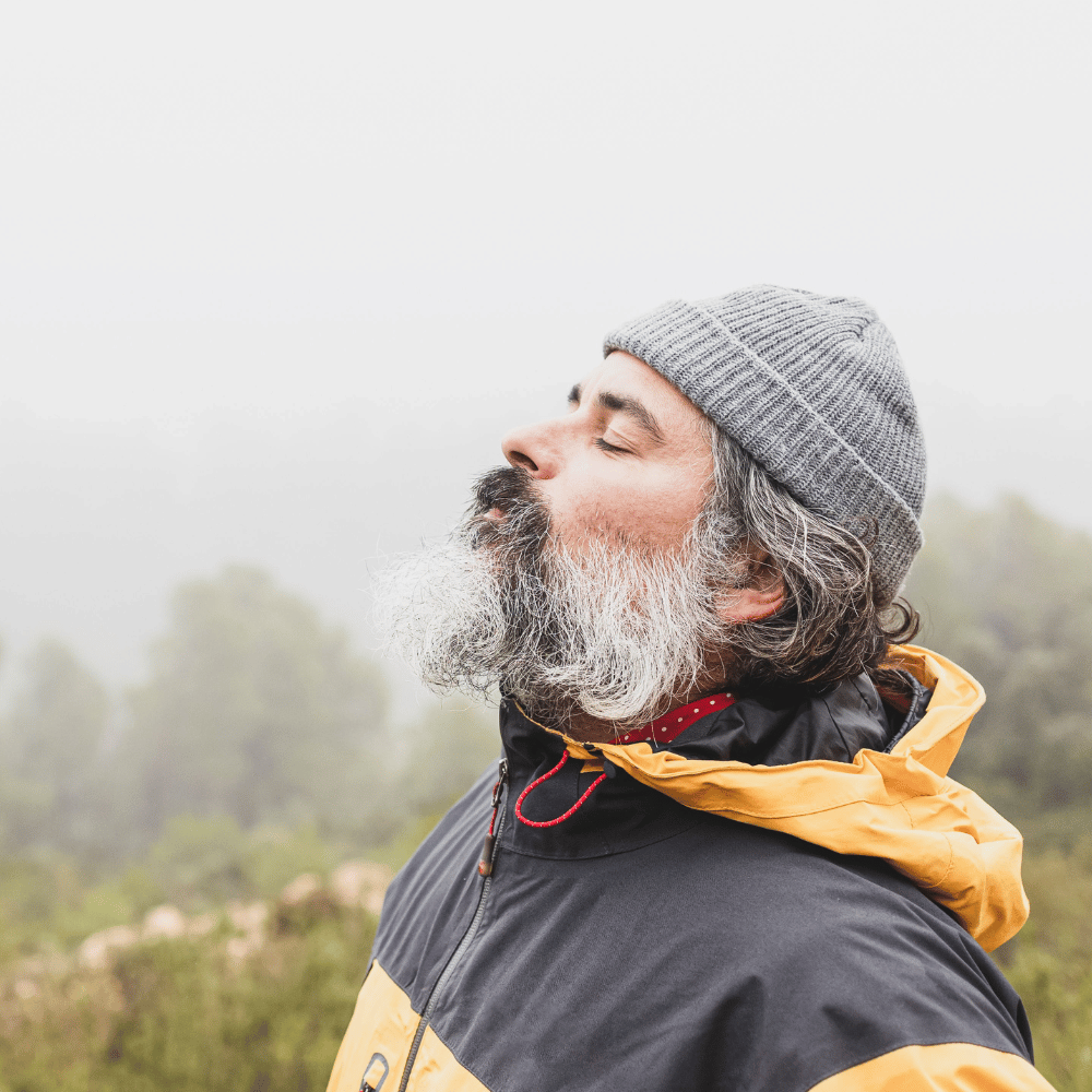 A bearded man in a gray and yellow jacket breathes deeply, representing outdoor men's apparel for an adventurous lifestyle.