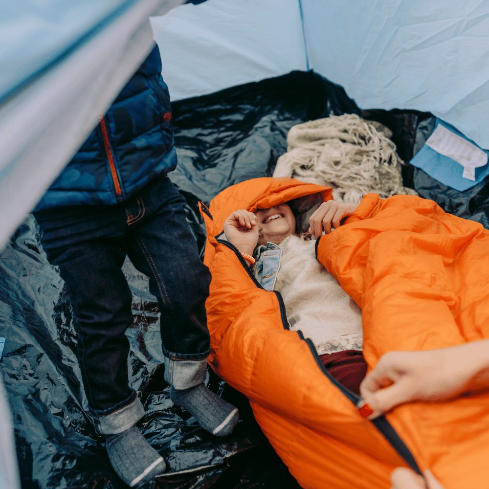 Inside a tent, a child in an orange sleeping bag laughs, beside a boy in jeans, evoking cozy, outdoor comfort.