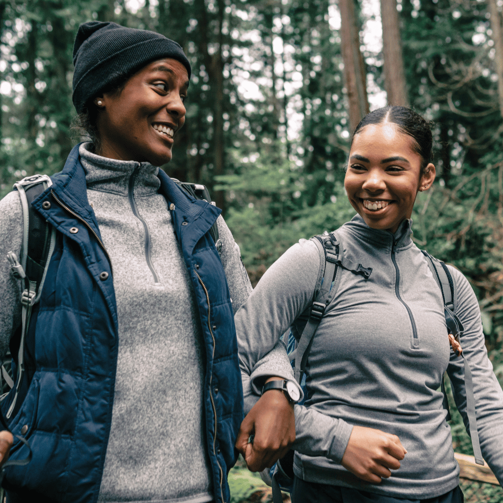 Two smiling women hiking in nature, wearing watches and backpacks, representing active, stylish accessories.