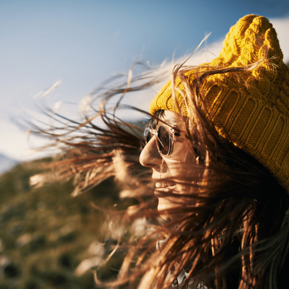 Person wearing a yellow knit hat with hair blowing in the wind against a blue sky.
