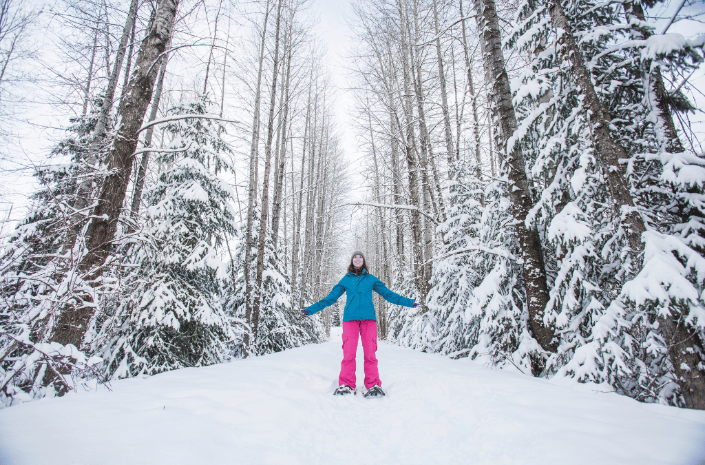 A woman in pink snow pants stands with open arms amidst snowy trees, conveying the joy of winter adventures and scoring affordable gear.
