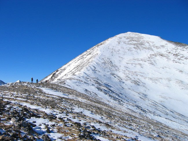 Hikers ascend a snow-covered fourteener under a clear blue sky, evoking a sense of challenge and adventure. 
