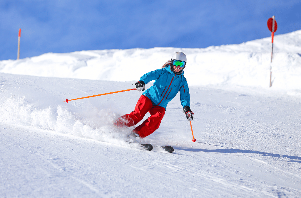 A skier in red pants and a blue jacket carves through fresh powder on a sunny slope.
