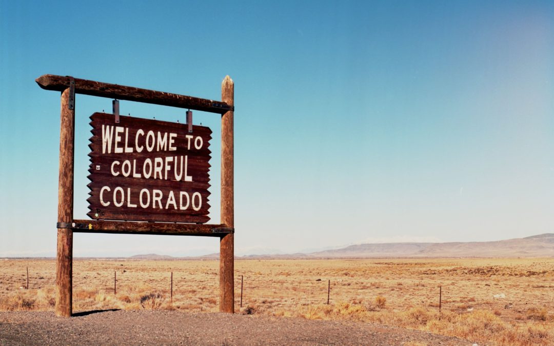 A weathered "Welcome to Colorful Colorado" sign stands in the vast, dry landscape, evoking a sense of adventure and stark beauty. 

