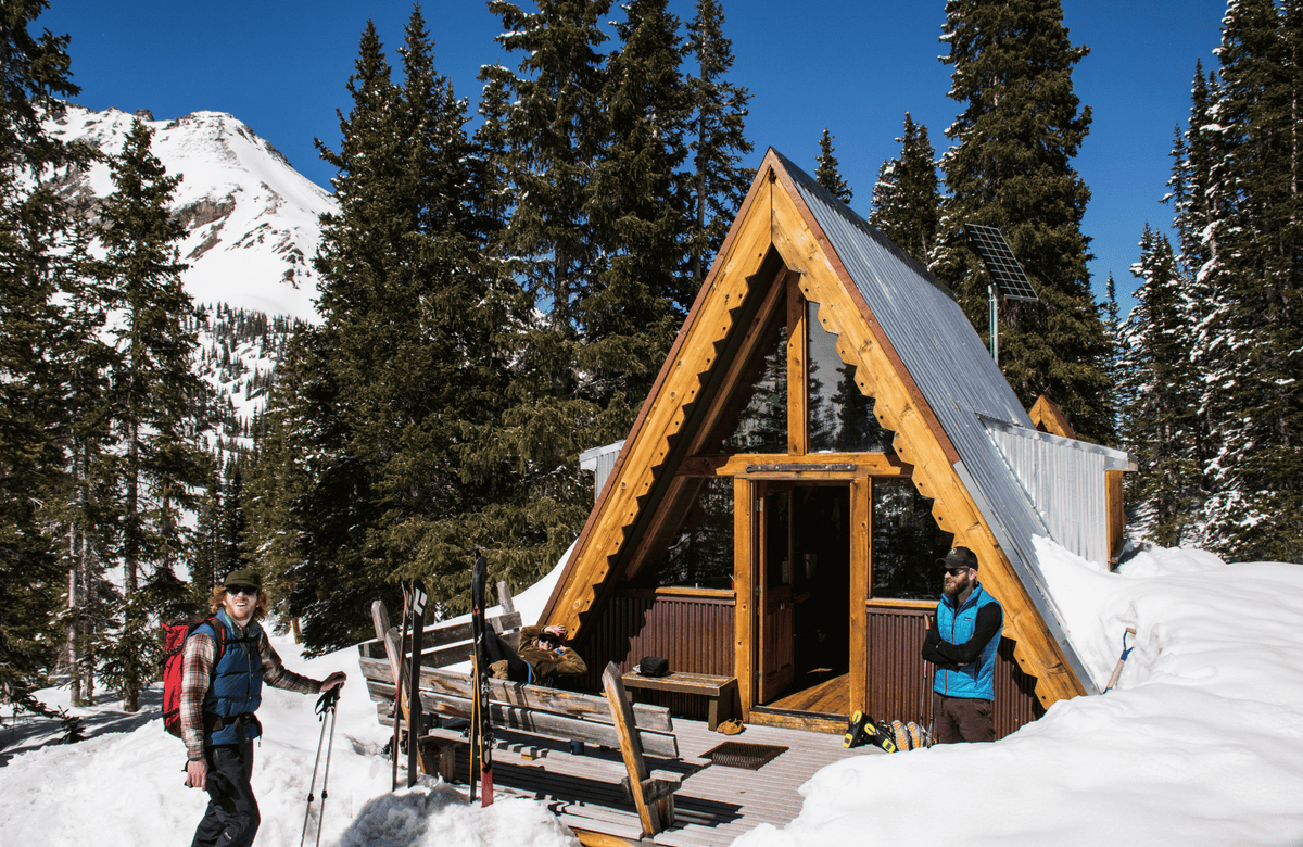 Snowy A-frame hut nestled in the mountains, inviting weary skiers for a cozy rest on their backcountry adventure.
