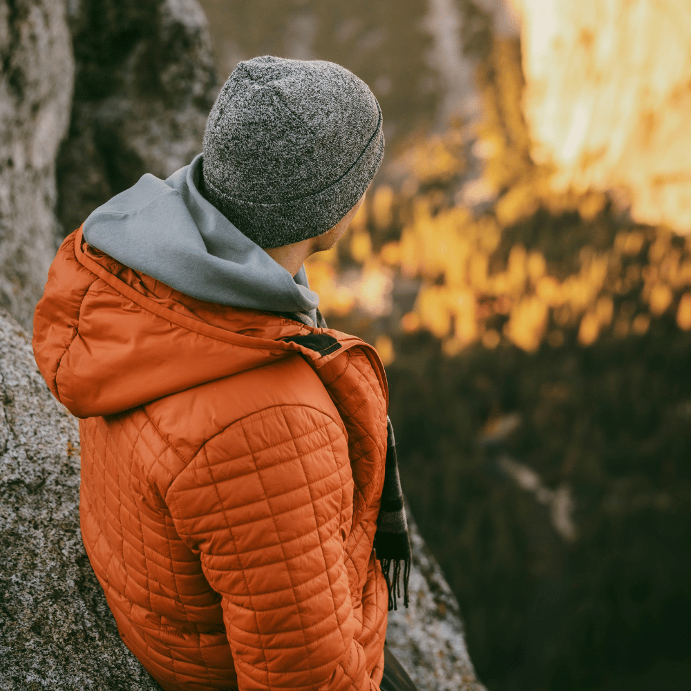Person wearing an orange quilted jacket and gray beanie standing in a natural setting with trees and rocks.