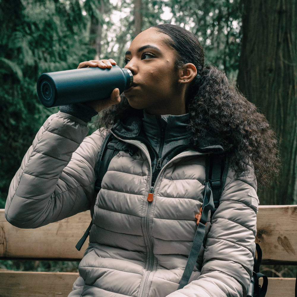 Woman in a puffer jacket drinking from a water bottle in a forest setting