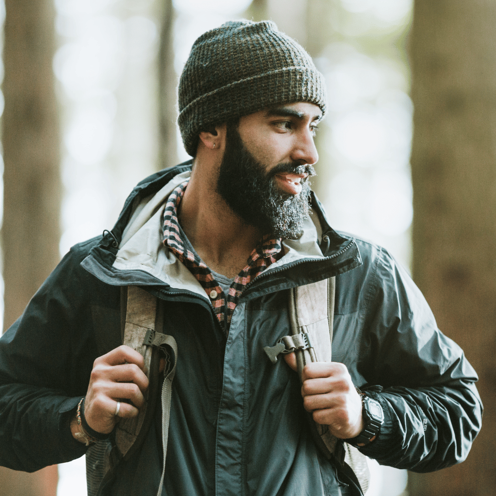 Man with a beard wearing a knit hat and jacket in a forest setting.