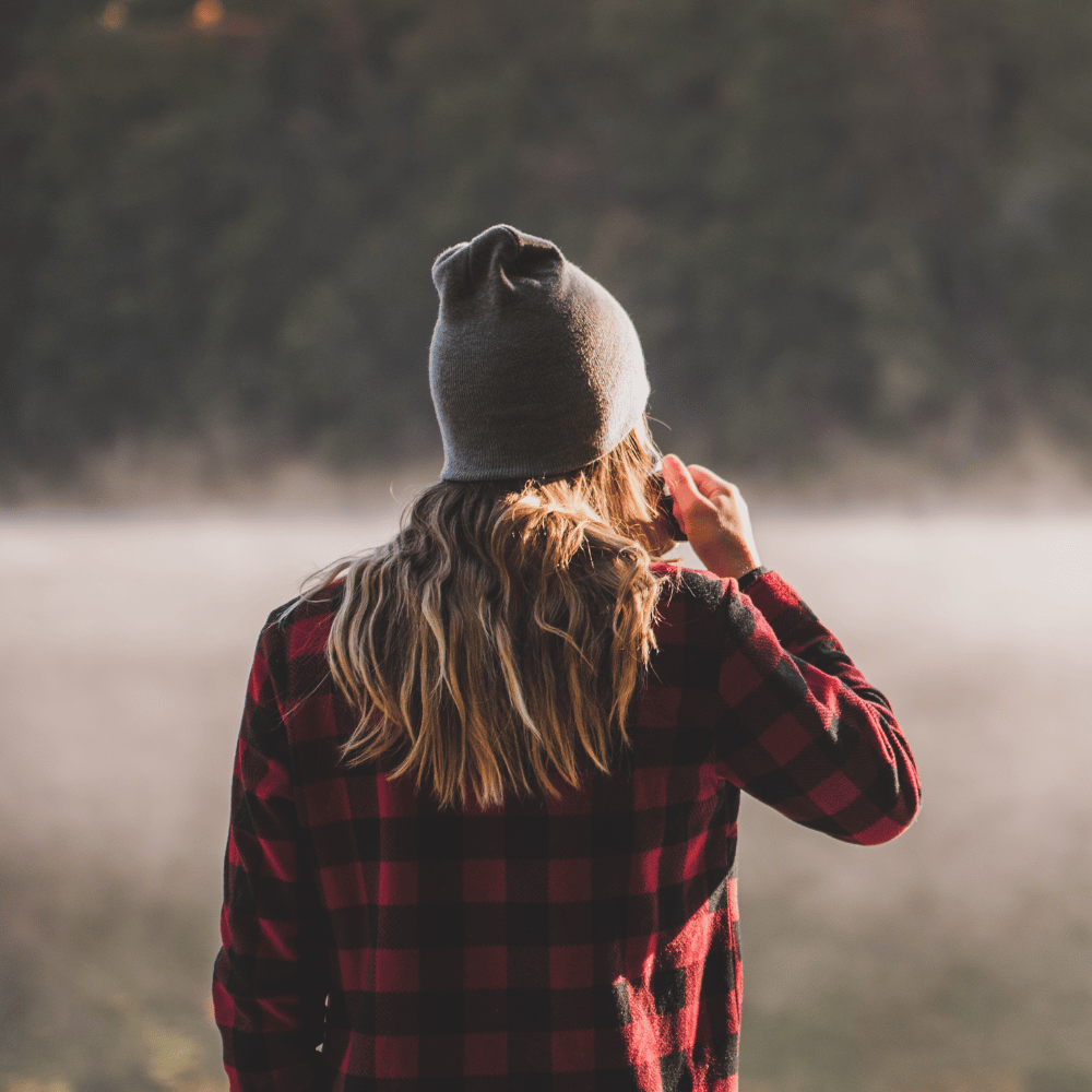 Person wearing a red plaid shirt and gray beanie standing by a misty lake.