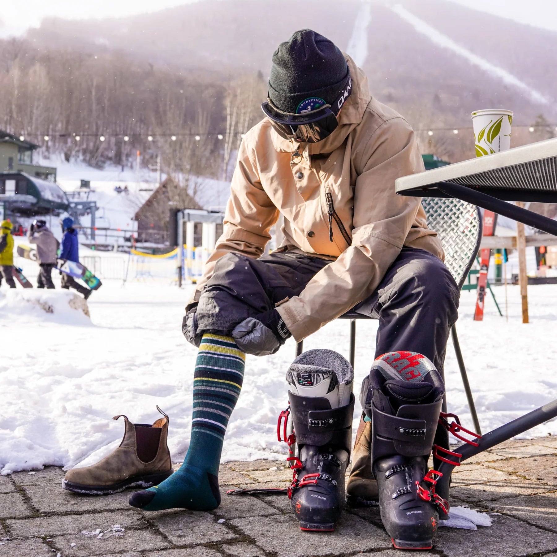 Striped ski socks by Darn Tough with yellow, white, green, and black lines, worn with boots in a snowy outdoor setting.
