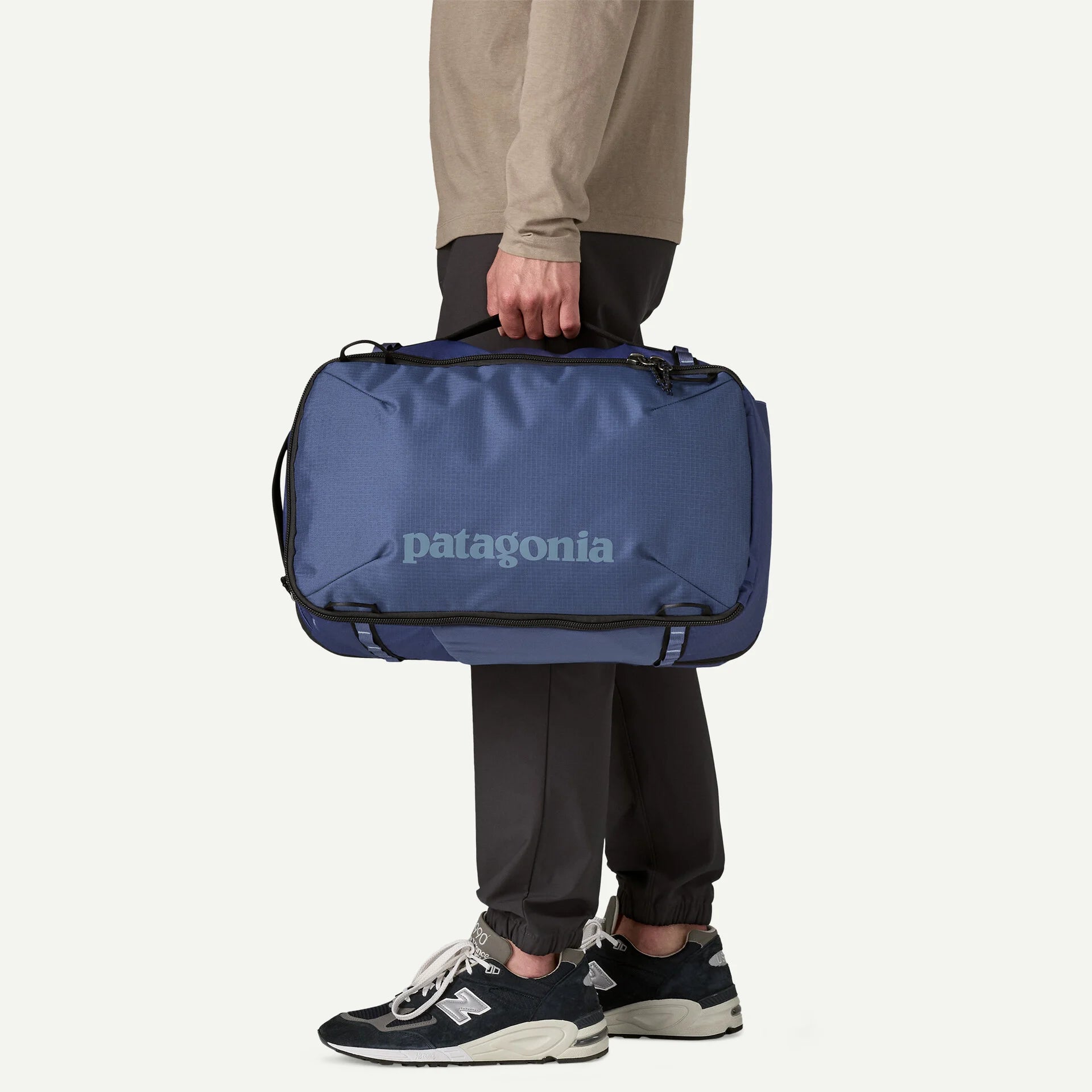 Blue travel backpack by Patagonia, seen held by the top handle, with neutral-colored shirt and dark pants and athletic shoes.