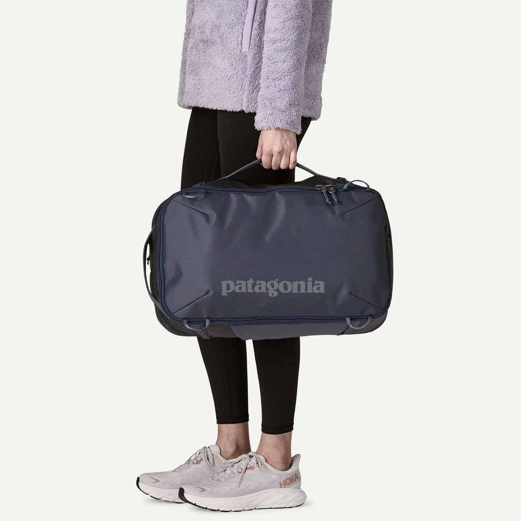 Navy travel backpack by Patagonia held in a studio setting, paired with a lavender fleece jacket and white sneakers.