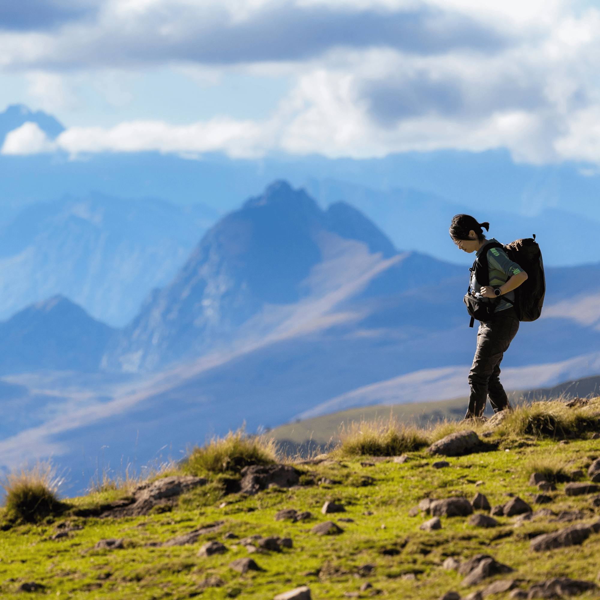 Hiker with a backpack standing on a grassy hill with mountains in the background