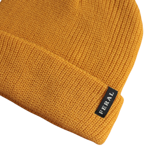 Golden knit beanie by FERAL, displaying ribbed texture and a folded brim with a branded tag on the left.