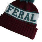 Burgundy knit beanie by FERAL with a white stripe featuring teal lettering, topped with a teal pompom.
