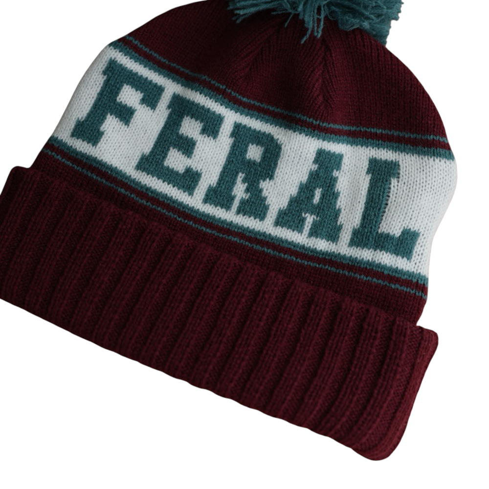 Burgundy knit beanie by FERAL with a white stripe featuring teal lettering, topped with a teal pompom.
