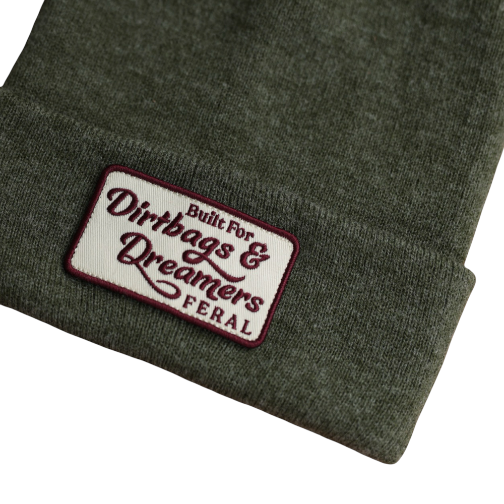 Olive green knit beanie by FERAL with a "Dirtbags & Dreamers" patch on the folded brim.
