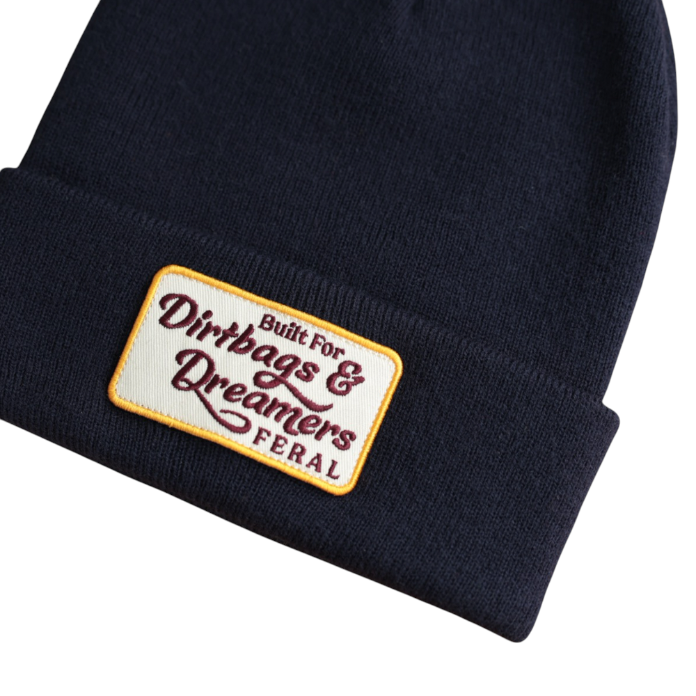 Dark blue knit beanie by FERAL, featuring an embroidered patch with the phrase "Dirtbags & Dreamers".
