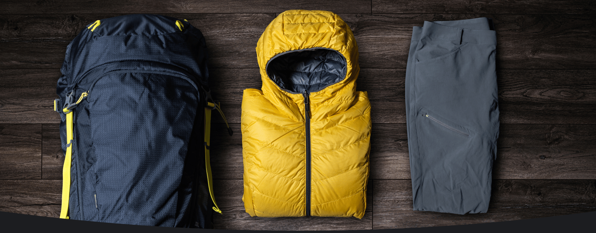 Three pieces of gear on a wooden surface: a dark blue backpack, a yellow puffer vest, and gray pants.