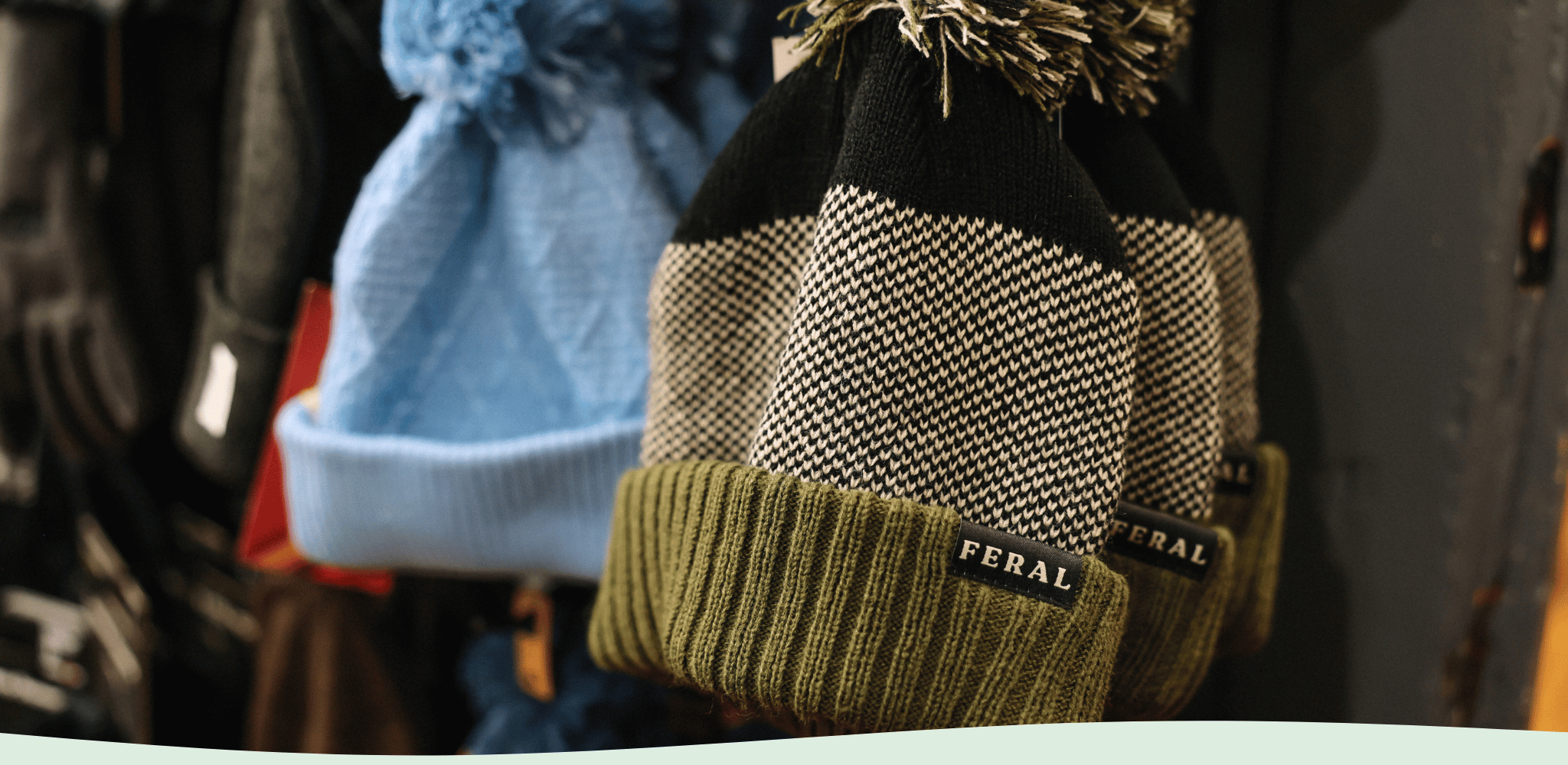 Green and Black FERAL Beanie Hanging on Wall Next to Blue Beanie