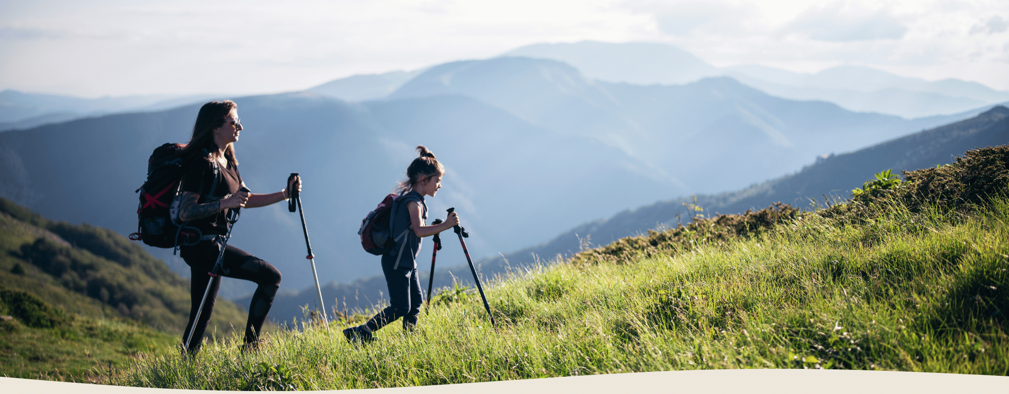 Two hikers with backpacks and walking sticks on a grassy hill with mountains in the background